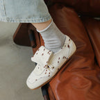 White sneakers with brown polka dots worn with gray socks on a brown leather chair.