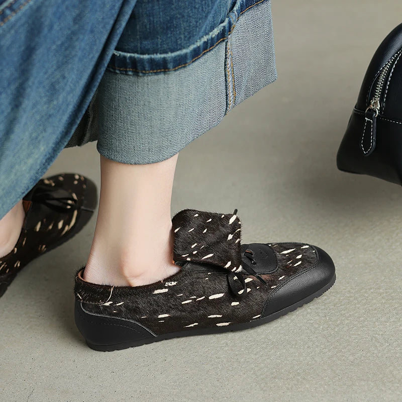 Black flat shoes with a bow detail worn by a person in jeans.
